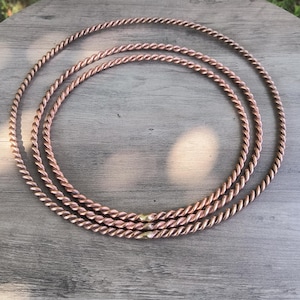 May include: Three copper-colored, twisted metal rings are arranged on a gray wooden surface. The rings are circular and have a textured, rope-like appearance. The background includes green grass and sunlight.