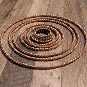 May include: Copper-colored, twisted metal rope arranged in concentric circles, creating a spiral pattern. The rope has a textured, braided appearance. The arrangement is on a wooden surface, with the rope's color contrasting the wood grain.