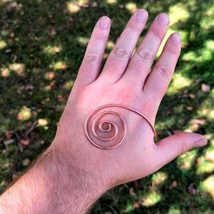 May include: A copper-coloured spiral-shaped bracelet. The bracelet is a continuous wire that forms a spiral design. The bracelet is resting on a hand, with a green and brown background.