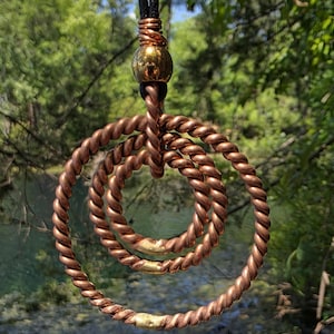 May include: A copper-colored pendant necklace with three intertwined, twisted rings. A gold-colored bead sits above the rings, connected to a black cord. The pendant hangs against a blurred background of green foliage and water.