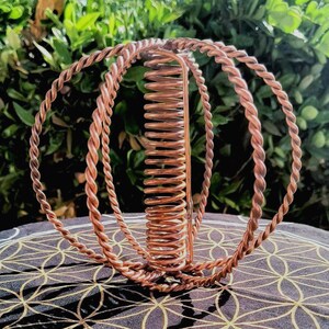 May include: A copper-colored metal sculpture with a spiral center and three surrounding twisted-wire rings. The sculpture sits on a patterned cloth with a green leafy background. The design is intricate and appears handmade.