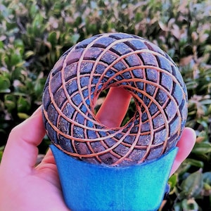 May include: A handcrafted, abstract sculpture with a unique design. The sculpture features a dark, textured, donut-shaped form with a woven pattern of copper-colored material. It rests on a blue base, creating a striking contrast. The artwork is held in a hand.