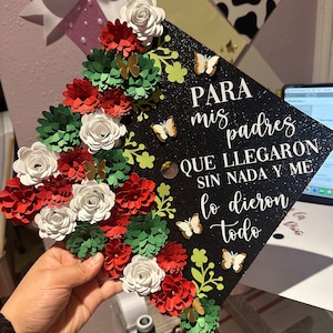 May include: A black graduation cap decorated with red, white, and green paper flowers and gold butterflies. The cap has the text "PARA mis padres QUE LLEGARON SIN NADA Y ME lo dieron todo" written on it.