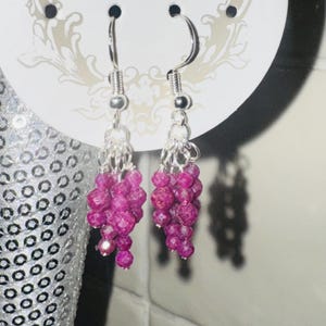 May include: Dangle earrings with clusters of small, magenta-coloured faceted beads. The beads hang from silver-toned hooks and chains. The earrings are displayed on a white card with a decorative design.
