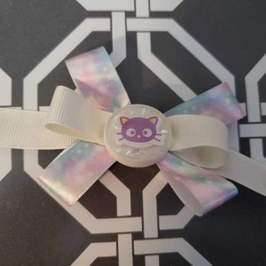 May include: A white and iridescent pink bow with a white ribbon and a purple cat face charm. The charm has a white background and a black outline.