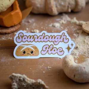 May include: A sticker with the words "Sourdough Hoe" in pink script, featuring a cartoon loaf of bread with a smiling face. The sticker is on a wooden surface with flour and baked goods.