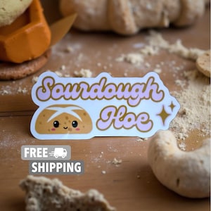 May include: A sticker with the text "Sourdough Hoe" in pink and gold lettering, featuring a cartoon bread loaf with a smiling face. The sticker is on a wooden surface with flour and baked goods.