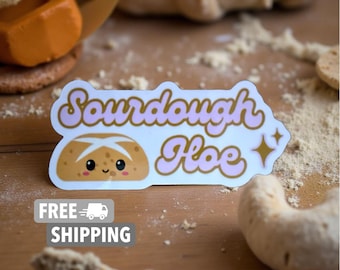 Op de afbeelding: Een sticker met de tekst "Sourdough Hoe" in roze en gouden letters, met een cartoon brood met een lachend gezicht. De sticker ligt op een houten oppervlak met bloem en bakkerijproducten.