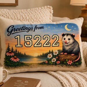 May include: Rectangular cushion with a scenic illustration. The design features a sunset over a lake, a smiling opossum, and the text "Greetings from 15222." Pink and white flowers and a crescent moon are also included.
