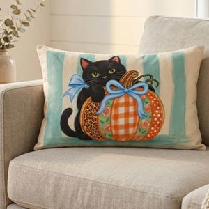 May include: A rectangular throw pillow featuring a black cat with a light blue bow sitting next to a pumpkin. The pumpkin has a checkered and leopard print pattern with a light blue bow. The background has vertical light blue and white stripes.