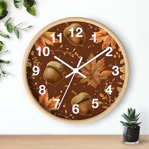 May include: Round wall clock with a light wood frame. The clock face features a brown background with illustrations of acorns and autumn leaves in shades of brown, orange, and gold. White numerals and clock hands complete the design.