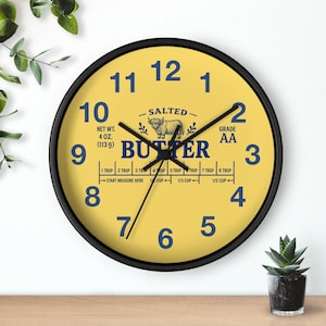 May include: A round wall clock with a yellow face and a black frame. The clock face features the word "BUTTER" and the text "NET WT. 4 OZ. (113 g)". The clock hands are black, and the numbers are blue.
