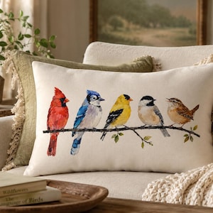 May include: A rectangular decorative pillow with a watercolor painting of five colorful birds perched on a branch. The birds include a red cardinal, a blue jay, a yellow finch, a black-capped chickadee, and a brown wren. The pillow is on a cream-colored background.