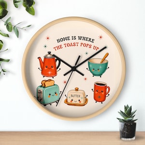 May include: A round wall clock with a light wood frame and a cream-colored face. The text reads "HOME IS WHERE THE TOAST POPS UP" above illustrations of a smiling orange kettle, a teal toaster, a butter dish, a teal bowl, and a red coffee mug.