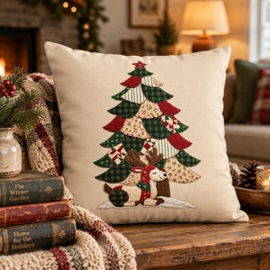 May include: A square beige throw pillow featuring a central applique design of a Christmas tree made from various patterned fabric patches in red, green, and cream, topped with a star, and a reindeer resting at the base.