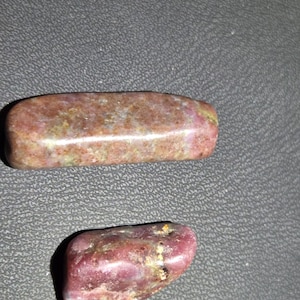 May include: Two polished, tumbled stones with a smooth, glossy texture. The larger stone is elongated and rectangular with rounded edges, featuring mottled shades of reddish-brown and green. The smaller stone is oval-shaped and deep magenta.