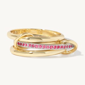 May include: A gold ring with an unusual interlocking design. The ring has a band of small, bright pink gemstones set between the gold bands. The ring is shown against a white background.