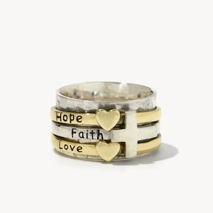 Peut inclure: Une bague tournante en argent et or avec les mots "Hope", "Faith" et "Love" gravés dessus. La bague comporte également deux petits cœurs en or et une croix en argent.
