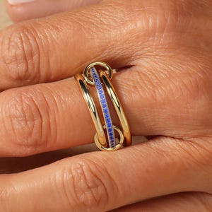 Pode incluir: Um anel de ouro com um design único, apresentando três bandas conectadas. Uma banda é adornada com uma fileira de pequenas pedras preciosas azuis. O anel é mostrado em um dedo, destacando seu estilo elegante e moderno.