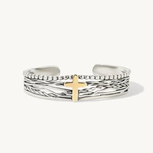 May include: Silver cuff bracelet with a gold cross in the center. The bracelet has a textured design with a beaded edge. The cross is centered and has a smooth, polished finish. The bracelet is open at the ends.