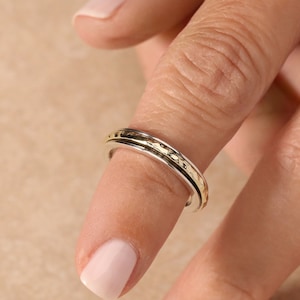 May include: A silver and gold spinning ring with a textured band. The ring is worn on a finger.