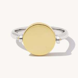 May include: A two-tone ring with a silver band and a large, round, gold-colored center. The ring has a modern design, with small gold accents where the center disc connects to the band. The ring is set against a white background.