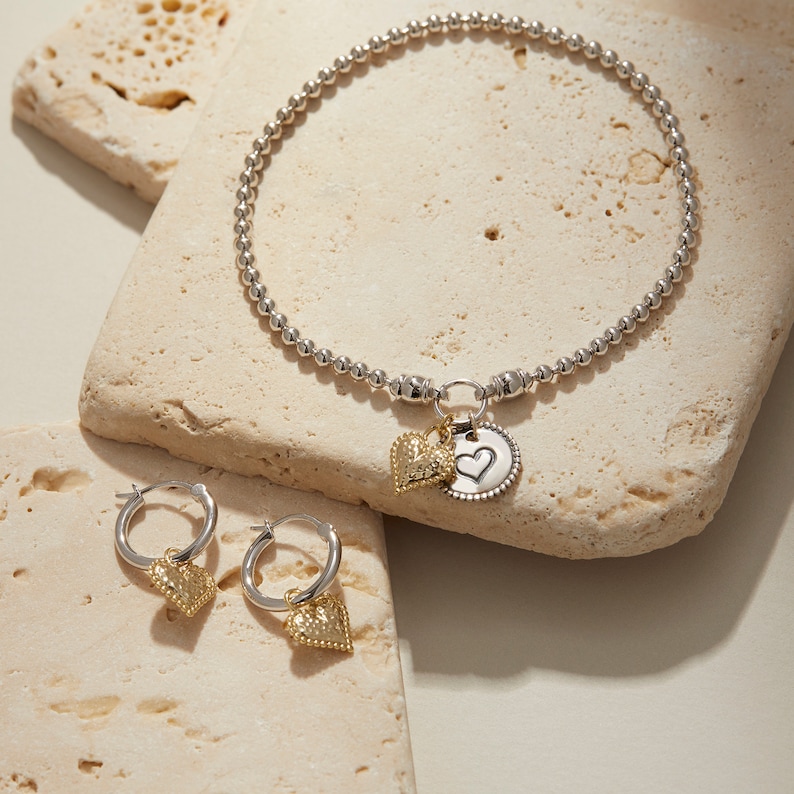 Puede incluir: Un collar de cuentas plateadas con un dije plateado con forma de coraz&oacute;n. El dije est&aacute; unido a un dije de coraz&oacute;n dorado m&aacute;s peque&ntilde;o. Tambi&eacute;n se incluyen dos aretes de aro de coraz&oacute;n dorados.