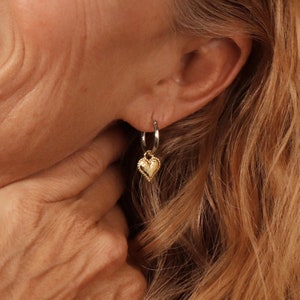 Puede incluir: Pendientes de aro de plata con un dije de coraz&oacute;n de oro. Los pendientes son usados por una persona con cabello largo y rubio.