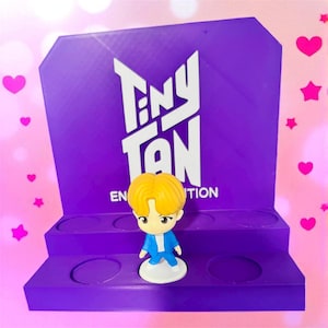 May include: A purple display stand featuring the text "TINY TAN" in white. A small figurine with yellow hair and a blue outfit stands on the stand. The background is decorated with pink hearts and stars.