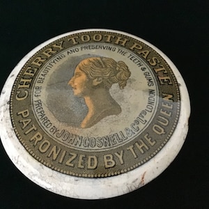 May include: A vintage, round, white ceramic container lid with a gold and brown design. The lid features a portrait of a woman with the text "Cherry Tooth Paste" and "Patronized by the Queen".
