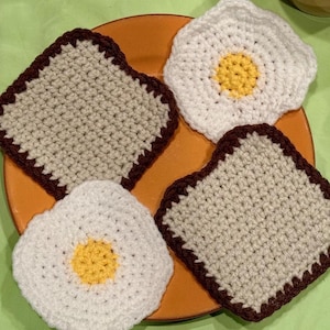 May include: Four crocheted coasters in the shape of toast and fried eggs. The coasters are brown, beige, and white.