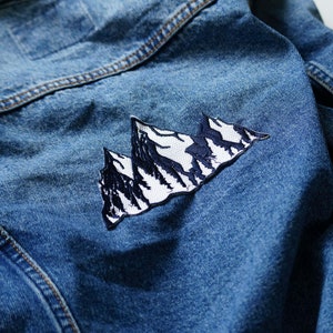 Peut inclure: Une veste en jean bleue avec un patch brodé de montagne blanc et noir au dos.