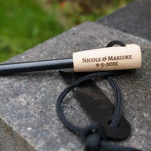 Könnte beinhalten: Ein schwarzer Feuerstarter mit einem Holzgriff, der mit "Nicole & Marieke 9-5-2025" graviert ist. Der Feuerstarter ist an einer schwarzen Schnur mit einem schwarzen Metallwerkzeug befestigt.