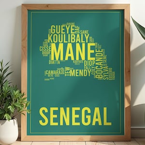 Op de afbeelding: Een ingelijste kunstdruk met een groene achtergrond en gele tekst. Het woord "SENEGAL" staat onderaan. Daarboven is een vorm te zien die is gemaakt van namen als "MANE" en "KOULIBALY". De lijst is gemaakt van lichtbruin hout.