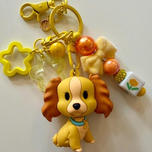 May include: A yellow dog-shaped keychain with brown ears and a blue collar. The keychain includes a star-shaped charm, a bow, and orange beads. The hardware is yellow.