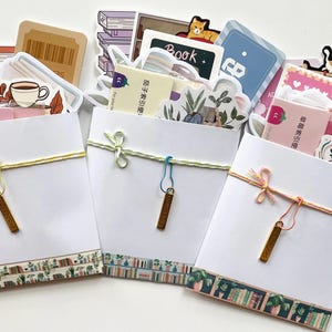 May include: Three white paper envelopes filled with assorted stationery stickers and cards. Each envelope is tied with colored string, featuring a small metal charm and a decorative border at the bottom depicting shelves of books and potted plants.