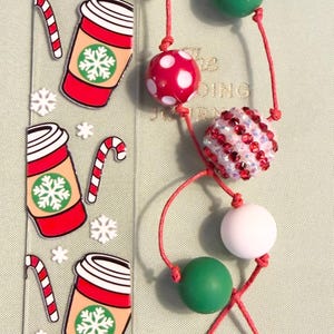 May include: A clear acrylic bookmark featuring printed graphics of red coffee cups with green snowflake logos and candy canes, accompanied by a red cord tassel adorned with white, green, red polka-dot, and red rhinestone-encrusted beads.