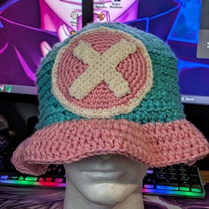 May include: A handmade crochet bucket hat in shades of teal, pink, and cream. The hat features a pink brim and a circular emblem with a cream-colored cross on a pink background. The hat is displayed on a white mannequin head.