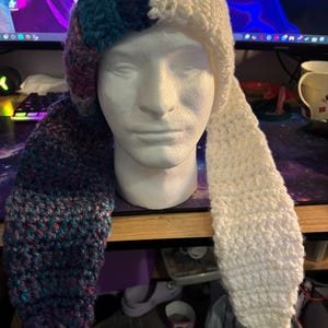 May include: A handmade crochet hat with long ear flaps. The hat features a color block design with sections of white, teal, and purple yarn. The hat is displayed on a white mannequin head, showcasing its unique style and craftsmanship.