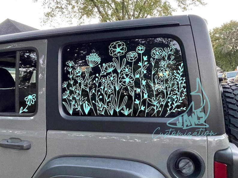 May include: A grey Jeep Wrangler with a teal floral decal on the rear window. The decal features a variety of flowers and leaves. The text "JAWS Customization" is visible on the lower right corner of the window.