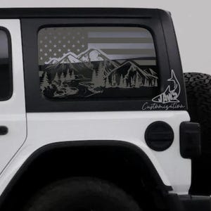 Puede incluir: Un Jeep Wrangler blanco con una calcomanía negra en la ventana trasera que representa una cordillera, un río y un bosque. La calcomanía también incluye una bandera estadounidense y el texto "JAWS Customization".