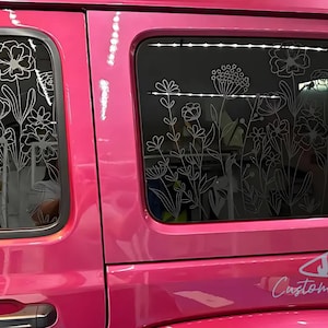 May include: A pink Jeep with a floral design etched into the rear windows. The Jeep has a shark fin decal on the back with the text "JAWS Customization" underneath.