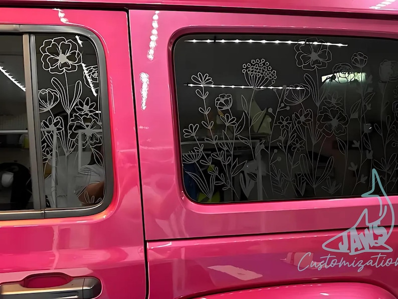 May include: A pink Jeep with a floral design etched into the rear windows. The Jeep has a shark fin decal on the back with the text "JAWS Customization" underneath.