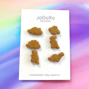 May include: A white card with the text "JoDoRo Designs" and "handmade clay jewelry" printed on it. Six brown clay dinosaur and cloud shapes are arranged on the card.