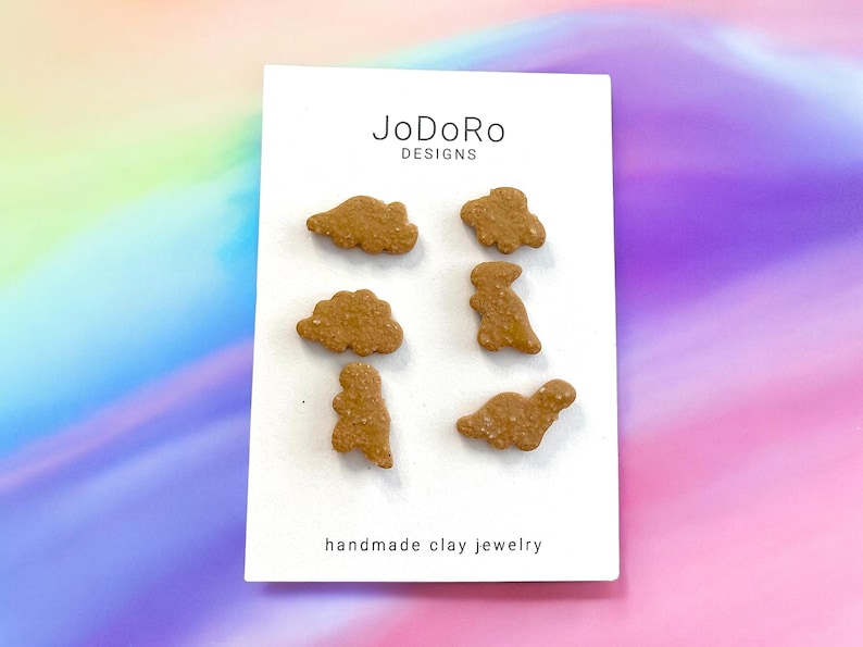 May include: A white card with the text "JoDoRo Designs" and "handmade clay jewelry" printed on it. Six brown clay dinosaur and cloud shapes are arranged on the card.