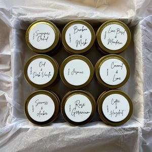 May include: A selection of nine small, gold-coloured tins, each with a white label and black text, presented in a box. The tins are labelled with various scents such as "Singapore Orchid", "Bamboo & Musk", and "Lime Basil & Mandarin."