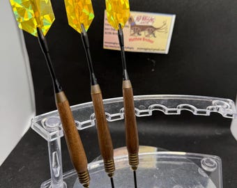May include: Three darts with yellow and iridescent flights are displayed in a clear acrylic holder. The darts have wooden barrels and black shafts. A business card is visible in the background.