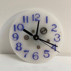 May include: A round clock face with blue numerals and black clock hands. The clock face is white with small, decorative gears. The clock hands point to 10:19. The clock face is a unique, decorative item.