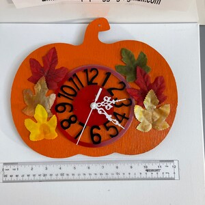 Op de afbeelding: Oranje pompoenvormige wandklok met een rode wijzerplaat en zwarte cijfers. De klok is versierd met kleurrijke kunstbladeren. De klok is ongeveer 30 cm lang.