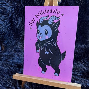 May include: A print featuring a cartoon goat with black fur, blue horns, and a blue face, standing on its hind legs. The background is a solid pink color with the words "live deliciously" in a curved font.